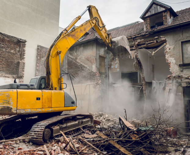 A yellow excavator tearing down an old, abandoned building, creating dust and rubble during demolition.